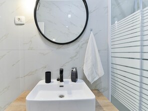 Un baño con un lavabo blanco, grifos negros, un espejo redondo y una toalla colgada en un perchero.