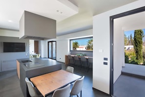 A modern kitchen with a central island, a dining area with a wooden table, and a view of the outdoors through large windows. - 3127 Villa Ottavia by Perle di Puglia (Porto Cesareo)