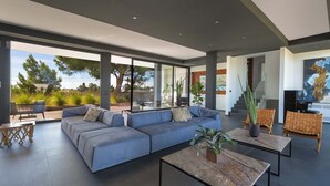 A modern living room with a large sectional sofa, a coffee table, and wicker chairs. There is a staircase leading to an upper level, and large windows providing a view of the outdoors. - 3127 Villa Ottavia by Perle di Puglia (Porto Cesareo)