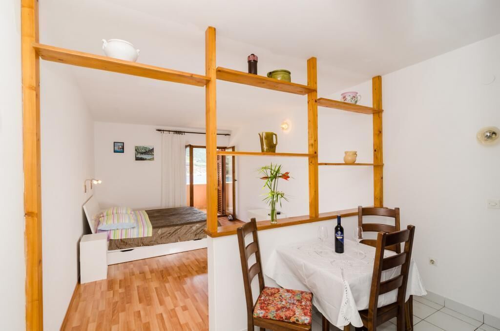 Una habitación con una cama, una mesa de comedor con sillas, un estante de madera con objetos decorativos y una planta pequeña.