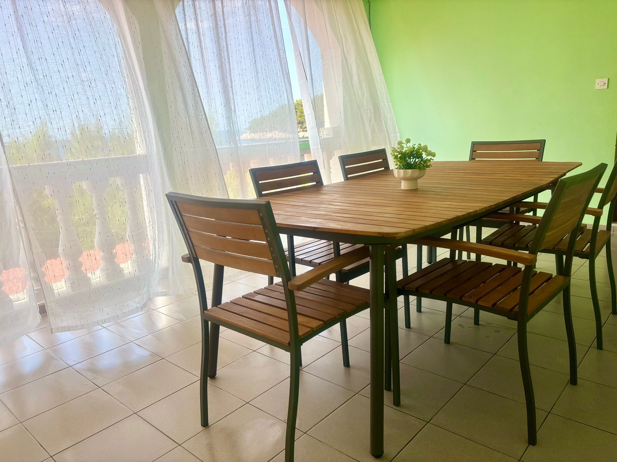 Comedor con mesa y sillas de madera, una planta en maceta y vistas a través de cortinas translúcidas.