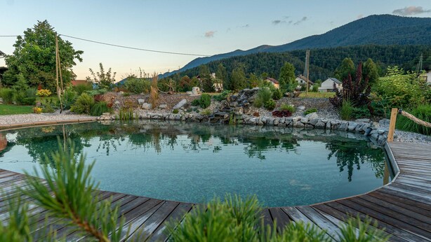 Ein Schwimmbad mit Holzterrasse, umgeben von Grünflächen und einer Bergkulisse.