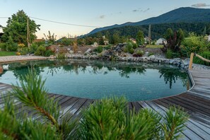 A swimming pool with a wooden deck, surrounded by greenery and a mountainous backdrop. - Cottage Happiness With Sauna and Natural Pool (Ribnica)