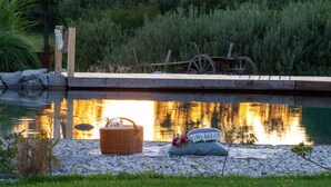 Ein Holzsteg erstreckt sich über einen stillen Teich, mit einer Picknick-Ecke, darunter ein Korb und Kissen, umgeben von Steinen und Grünpflanzen.