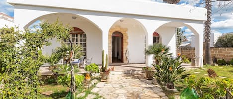 A white building with three arches, a stone pathway, and various potted plants.
