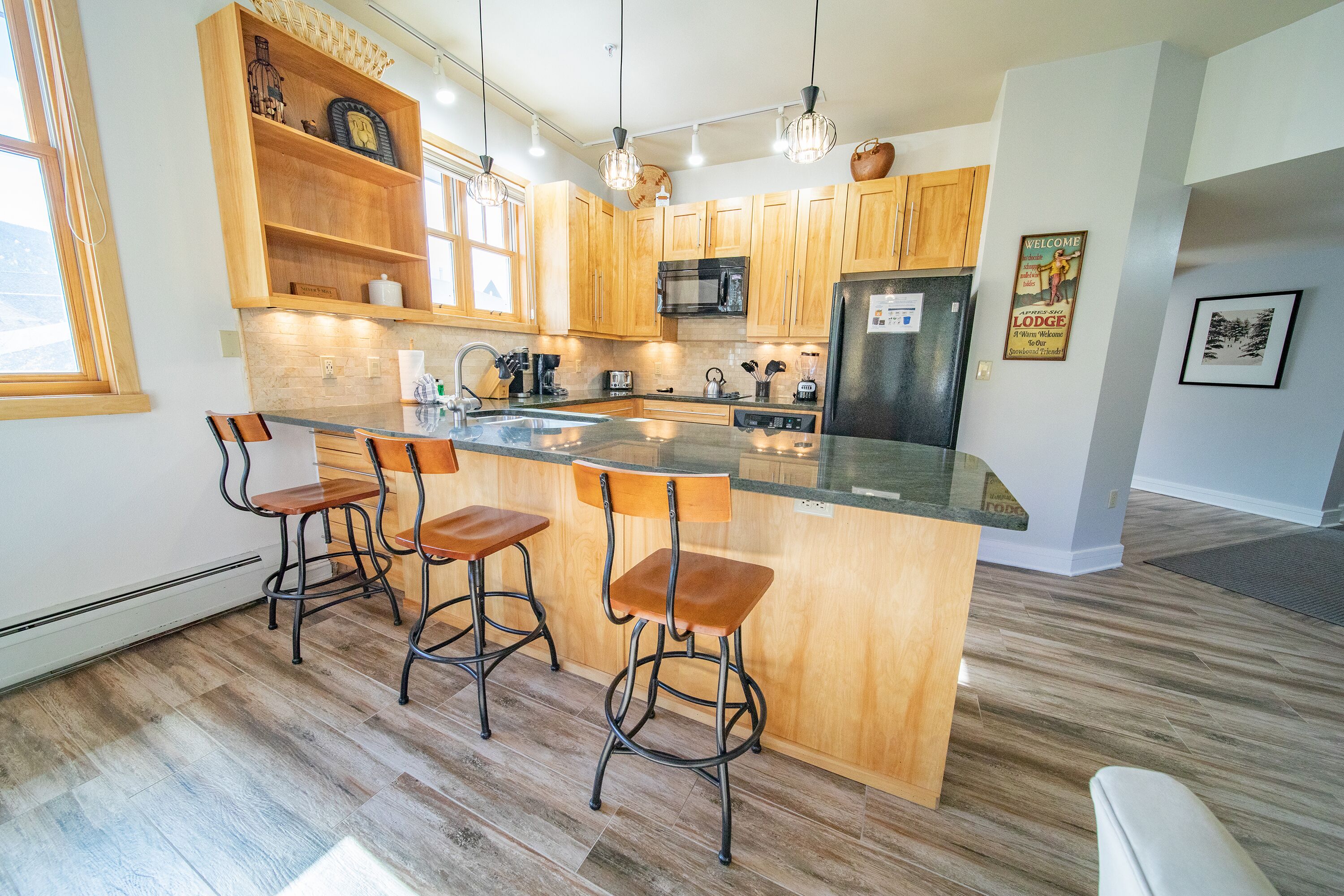 A modern kitchen with wooden cabinets, a central island with bar stools, a stainless steel refrigerator, and a microwave.