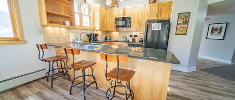 A modern kitchen with wooden cabinets, a central island with bar stools, a stainless steel refrigerator, and a microwave.