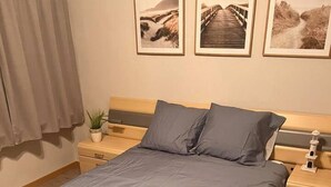 A neatly made bed with a grey bedspread and pillows, a wooden headboard, a nightstand with a lamp, and a small potted plant. - Beautiful 2-bed Apartment in Wenduine (De Haan)