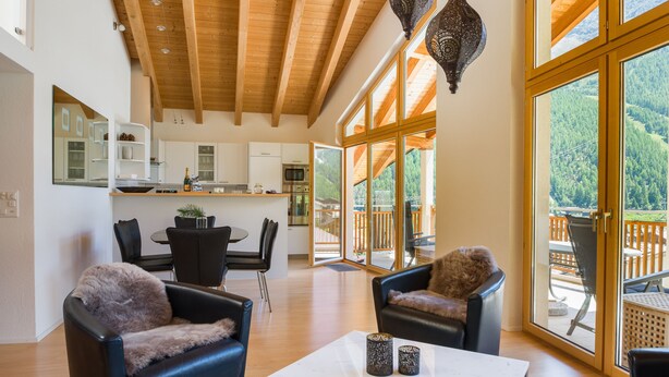 A spacious living area with a high wooden ceiling, large windows offering a view of the outdoors, and a modern kitchen in the background. - Impeccable 3-bed Duplex Penthouse in Saas-fee (Saas-Fee)