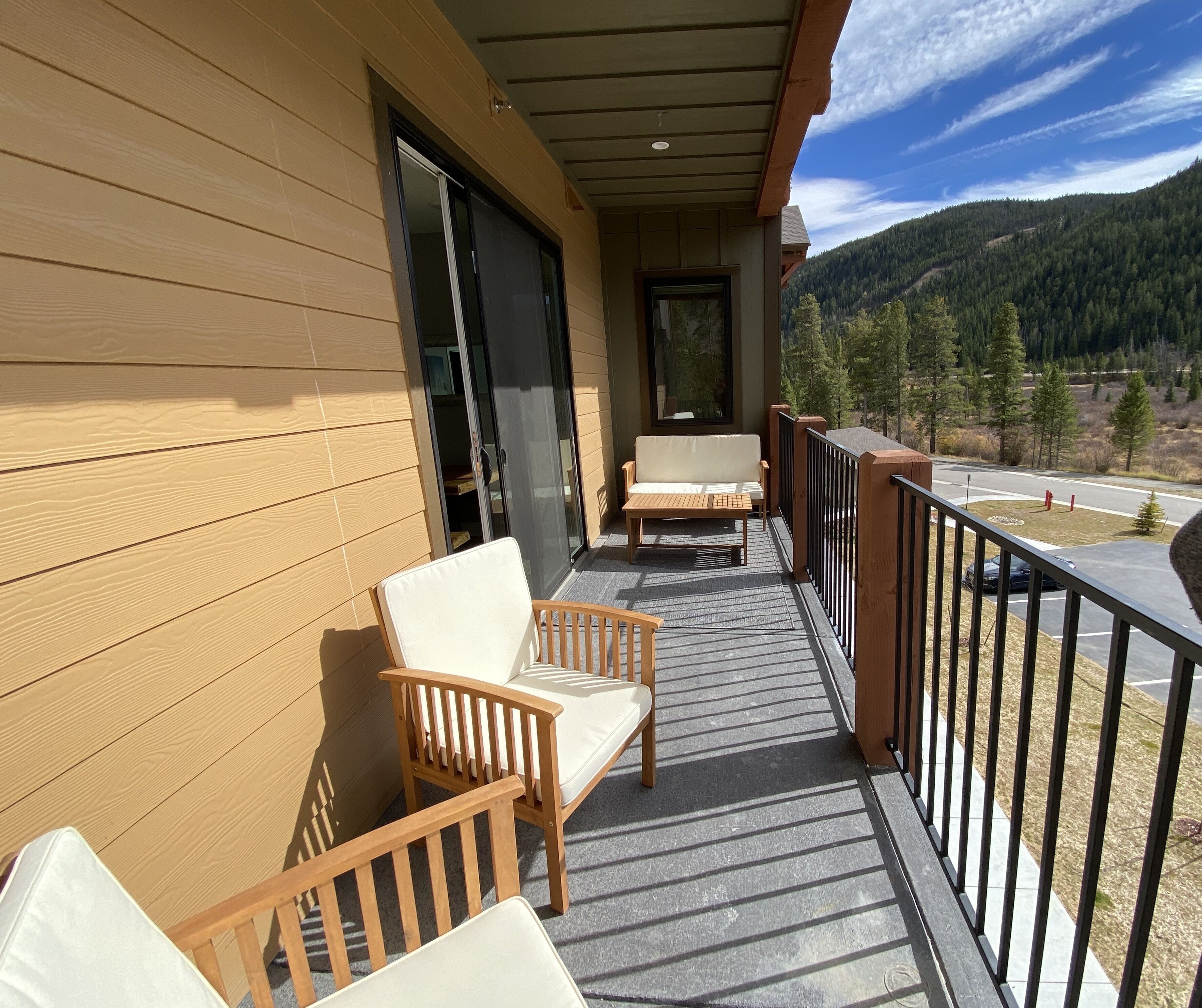 A wooden balcony with two chairs and a bench, overlooking a road and forested hills.