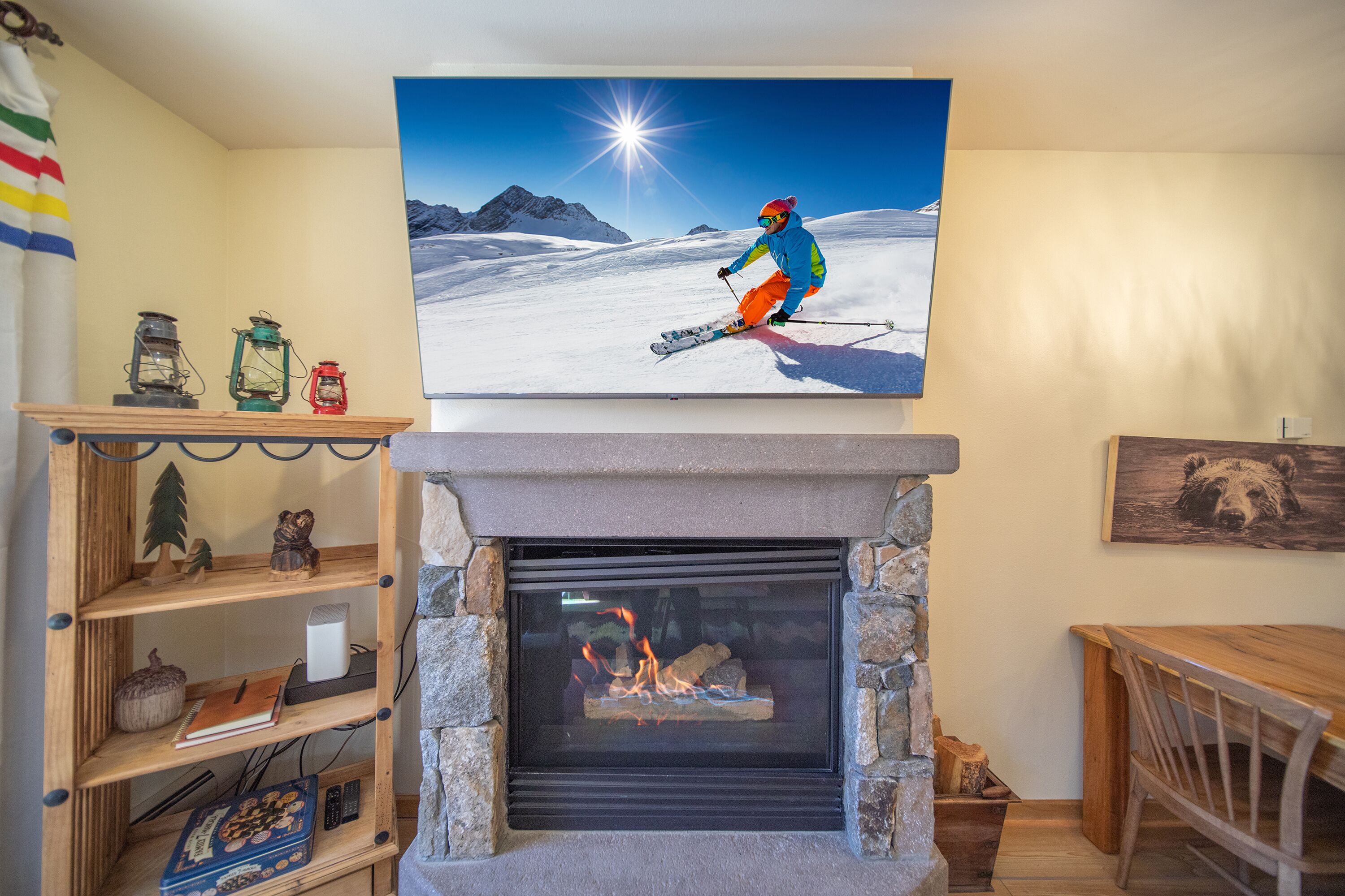 A fireplace with a stone surround, a wooden shelf with books and a small tree, and a painting of a skier above the fireplace.