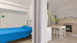 A bedroom with a single bed, a small bedside table, and a nightstand. The room has white walls and a window with a view of the outside.