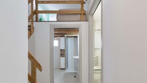 A narrow hallway with a wooden staircase on the left, leading to a room with a white sink and a glass shower enclosure.