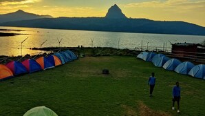 Un terrain de camping avec des tentes disposées en rangées, un champ d’herbe et une vue sur une montagne et un lac au coucher du soleil.