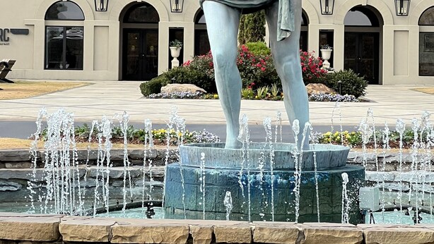 Una estatua de una mujer con vestido, de pie sobre una fuente frente a un edificio con una bandera, ventanas arqueadas y un letrero que dice "Restaurant".