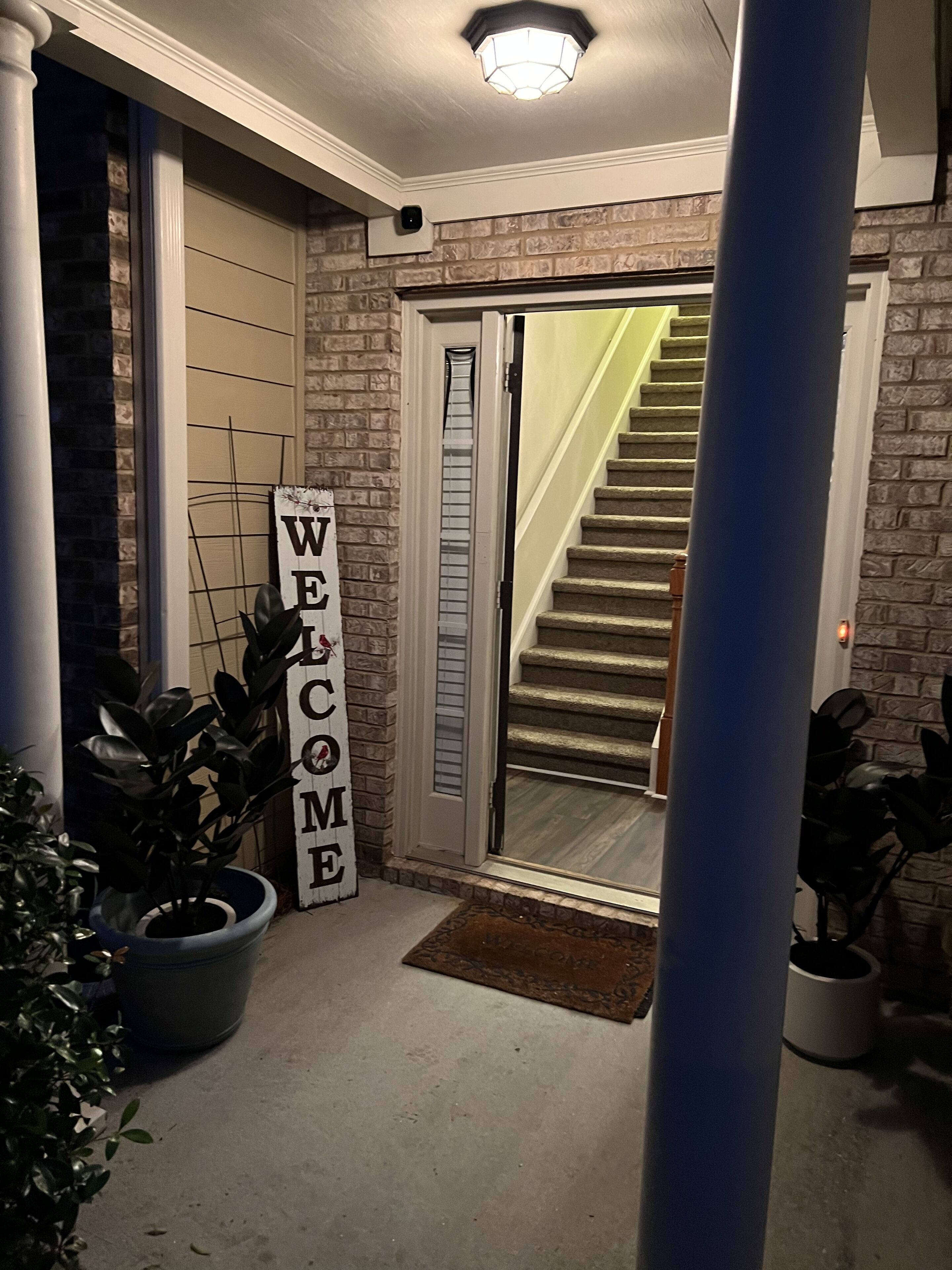 A staircase leading to a door with a 'WELCOME' sign, a potted plant, and a light fixture.