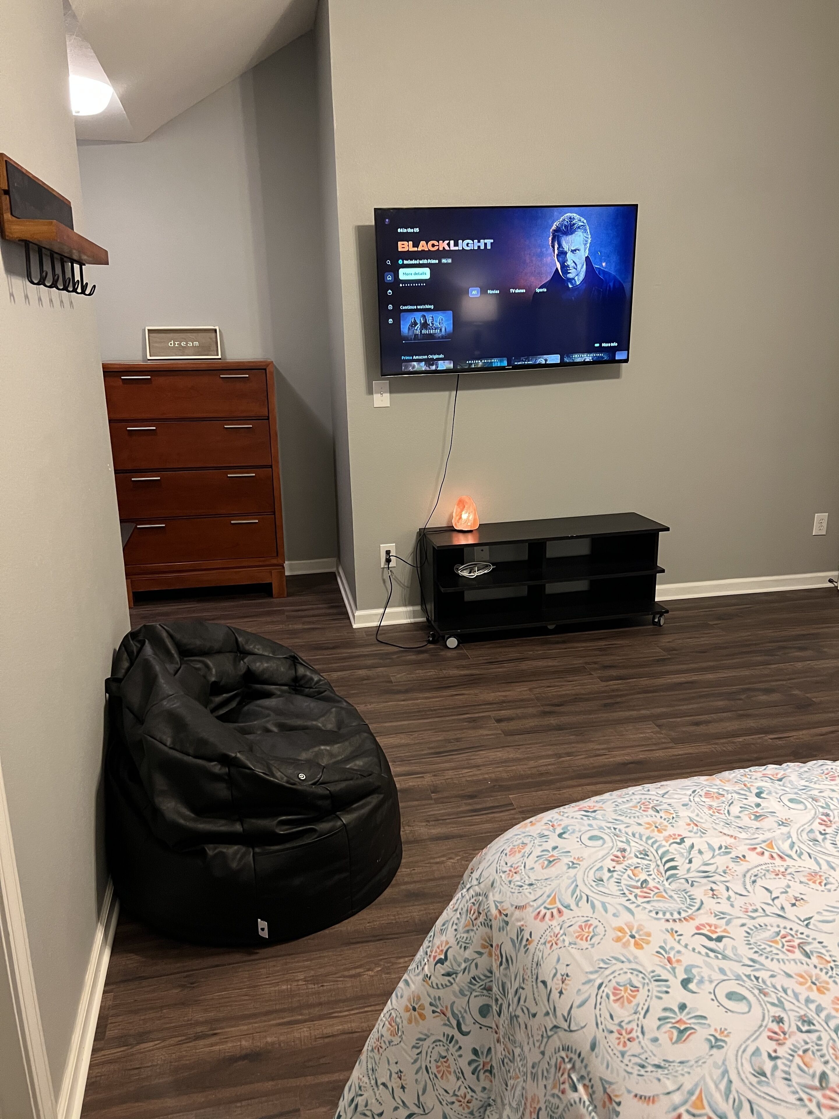 A bedroom with a bed, a black bean bag, a TV mounted on the wall, a wooden dresser, and a small table with a lamp.