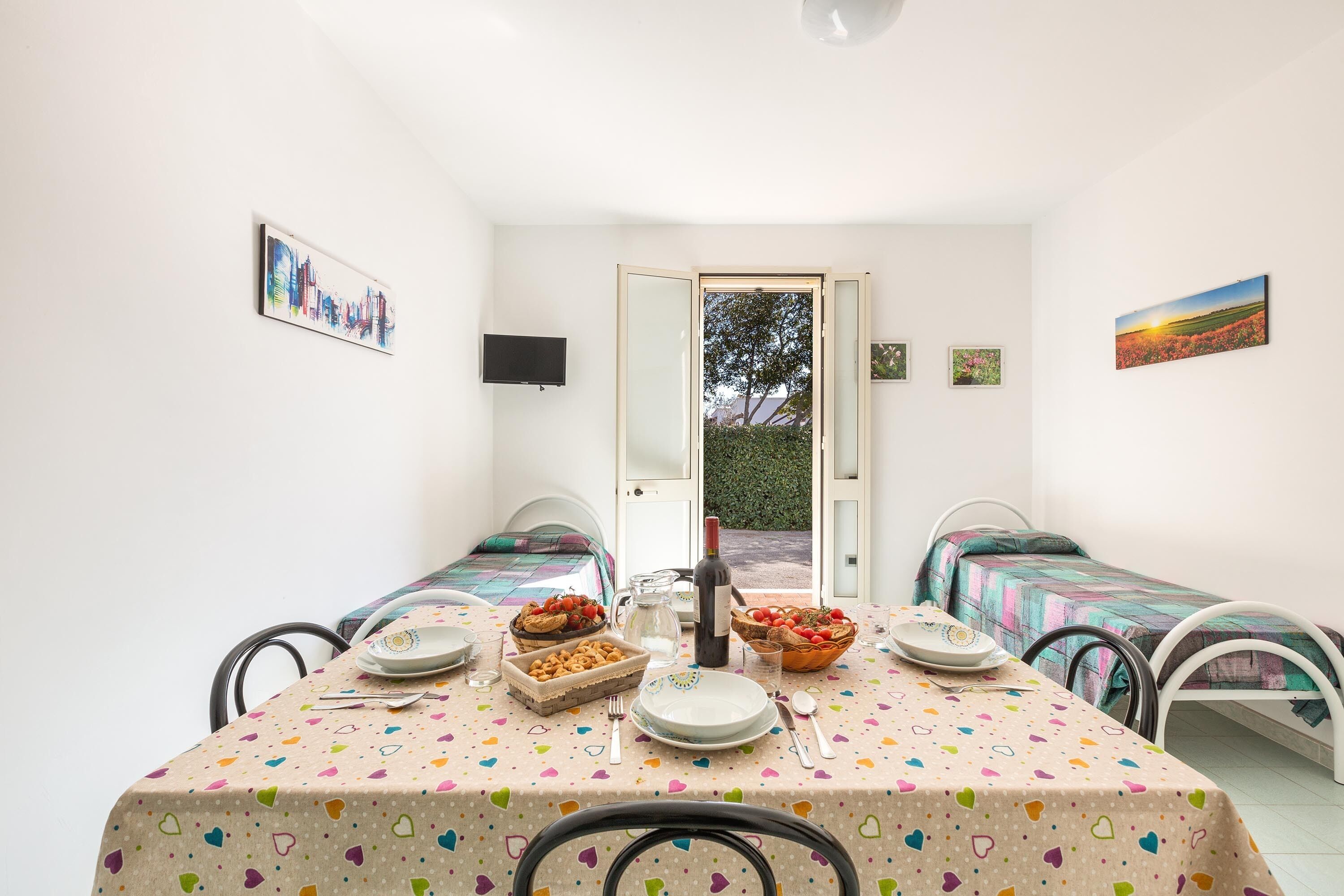 A cozy room with two single beds, a dining table set with plates and a bottle, and a TV mounted on the wall.