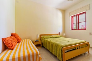 A room with two beds, one with orange and white striped bedding and the other with green bedding. There is a window with red frames and a view of greenery outside.
