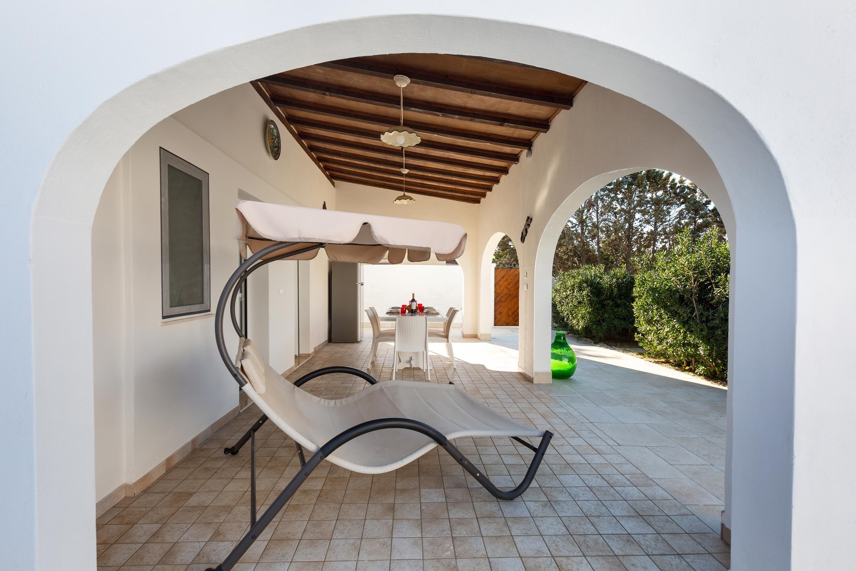 A covered patio area with a reclining chair, a table with chairs, and a view of the surrounding greenery.