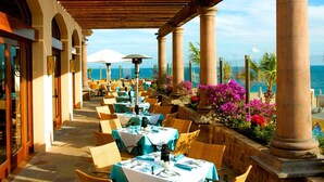 Un espace extérieur abrité, aménagé pour les repas, avec des tables, des chaises, des colonnes et une vue sur la mer.