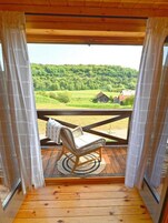 Una terraza de madera con una silla de mimbre y una pequeña mesa, con vistas a un paisaje verde que incluye un granero.