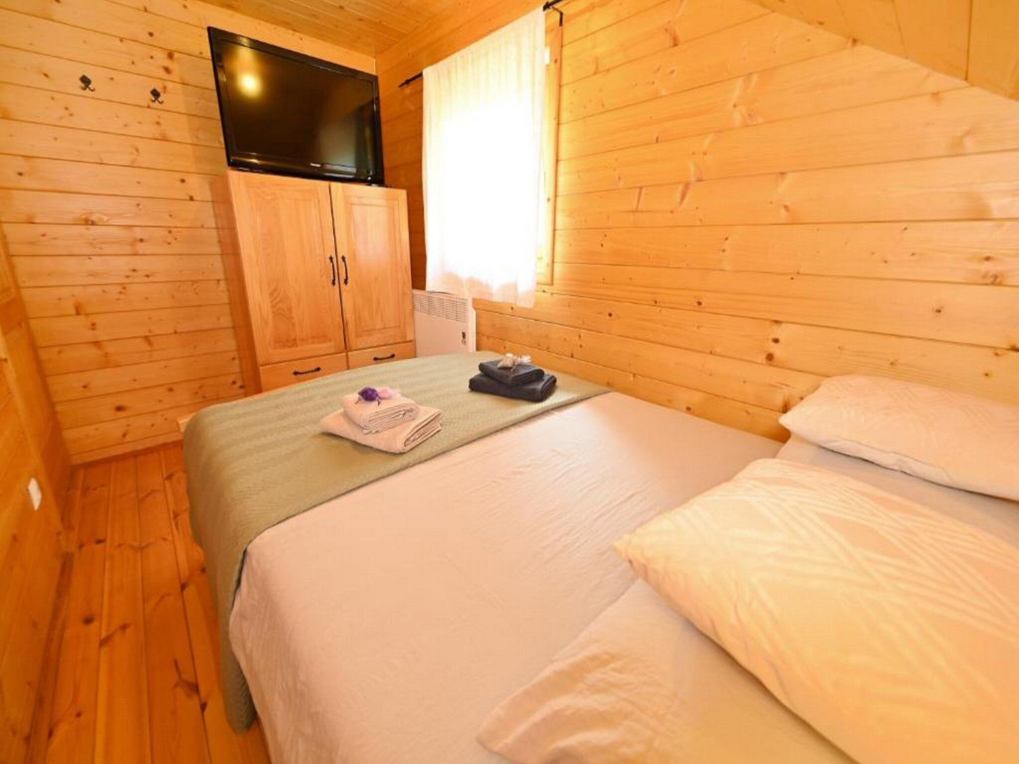 A wooden cabin room with a bed, a TV on a cabinet, and a window with curtains.