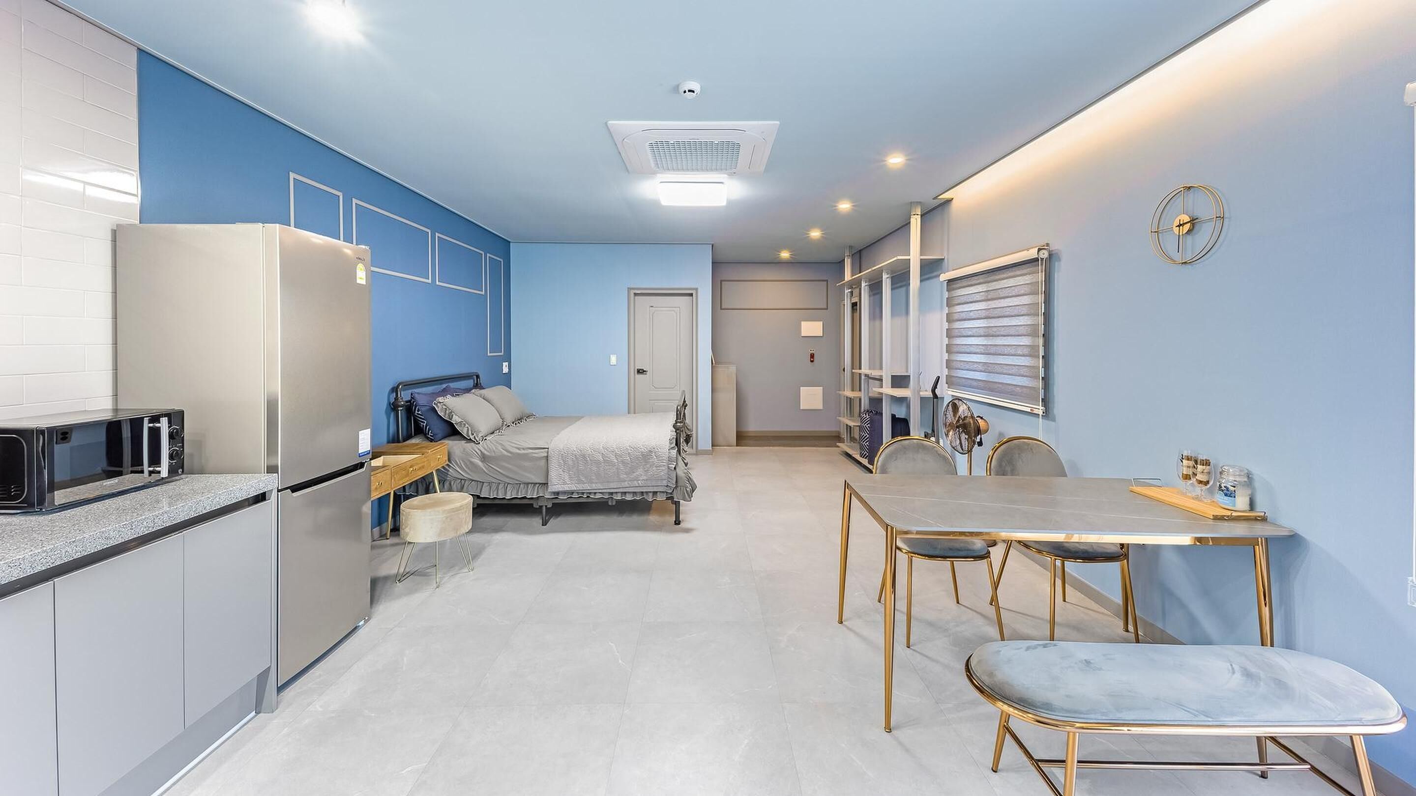 Cozinha compacta com geladeira, micro-ondas e torradeira. Uma cama com cabeceira e travesseiros no centro. Uma mesa de jantar com cadeiras à direita.