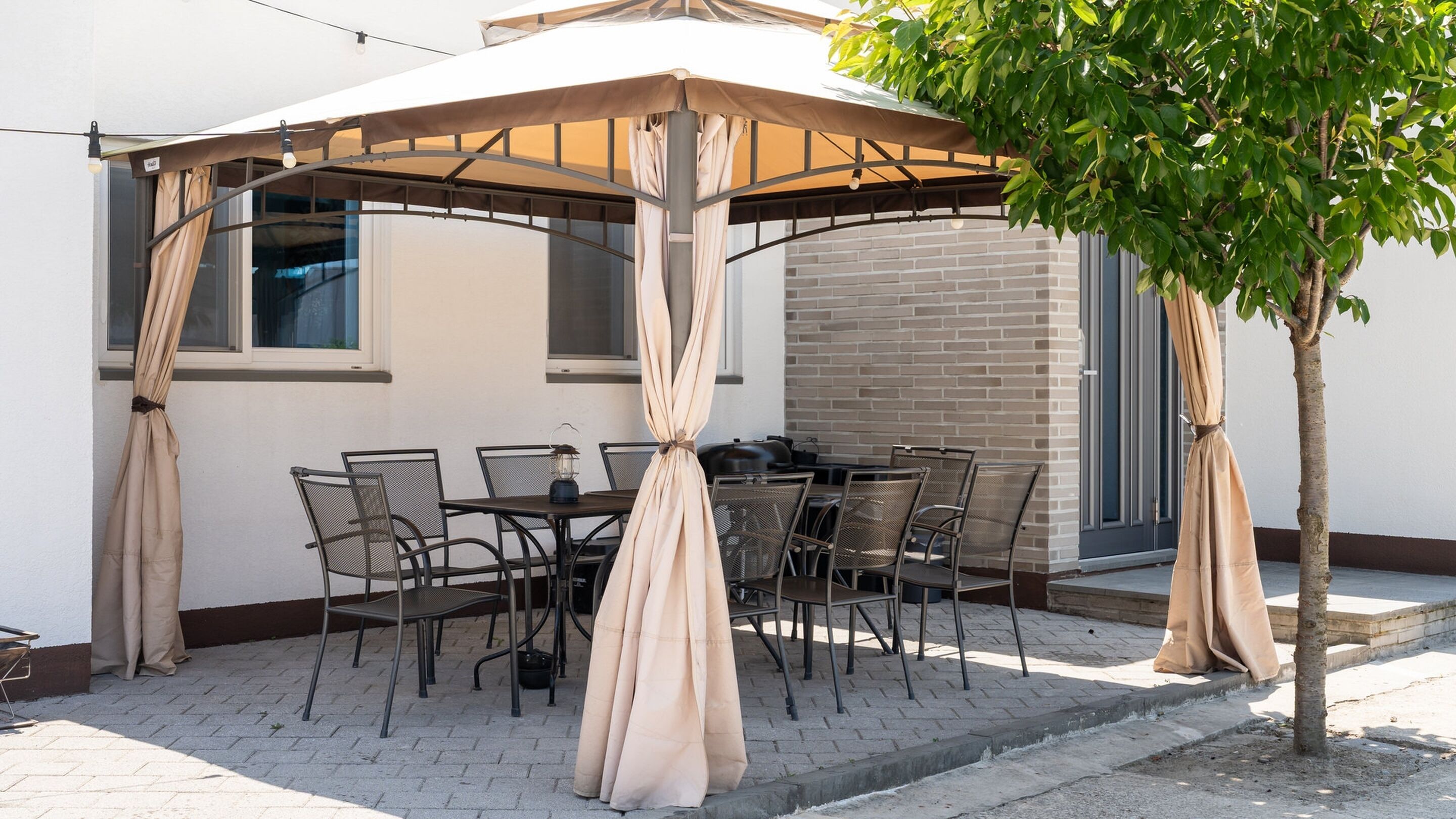 An outdoor patio with a covered table and chairs, a small tree, and a brick wall in the background.