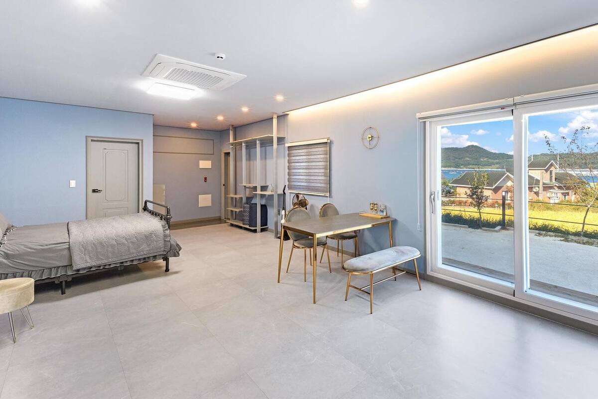 A bedroom with a bed, a desk, and a chair, a bathroom with a shower, and a large window with a view of a house and landscape.