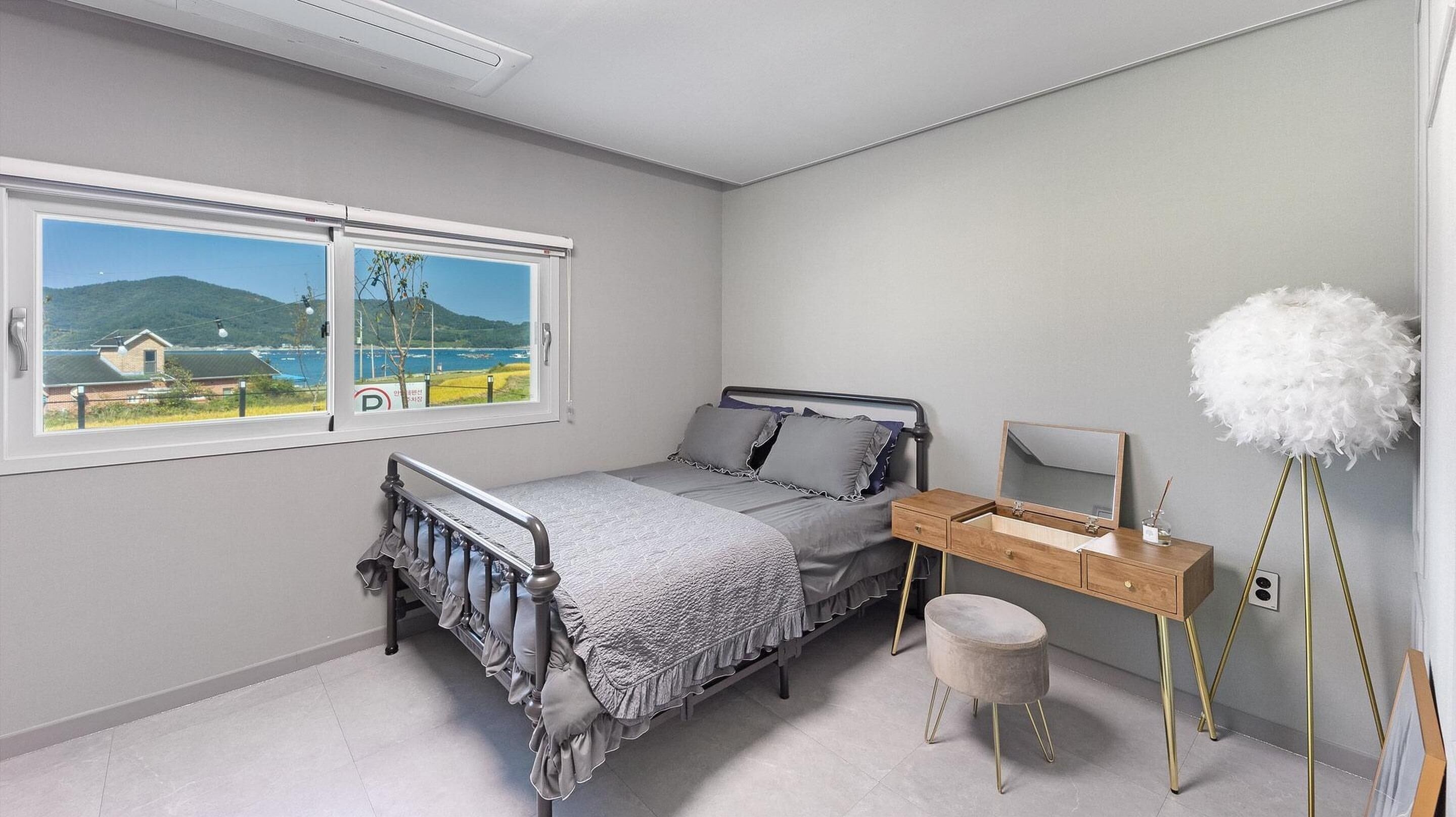 A bedroom with a bed, a desk with a mirror, and a view of a coastal landscape through a window.