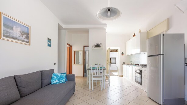 A modern living room with a grey sofa, a dining table, and a kitchen area with a refrigerator.