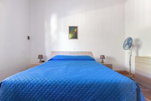 Una habitación con una cama, un cuadro colgado en la pared, un ventilador de pie y lámparas de noche.