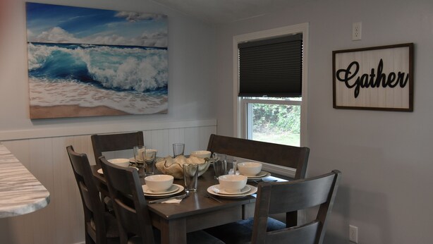Uma sala de jantar com mesa posta para quatro pessoas, uma pintura grande de uma onda na parede e um letreiro com a palavra 'Gather'.