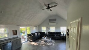 Uma sala de estar ampla, com ventilador de teto, sofá de couro escuro, mesa de centro branca e uma área de cozinha com forno embutido.