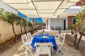 A covered outdoor dining area with a table set for four, surrounded by white plastic chairs, under a white canopy.