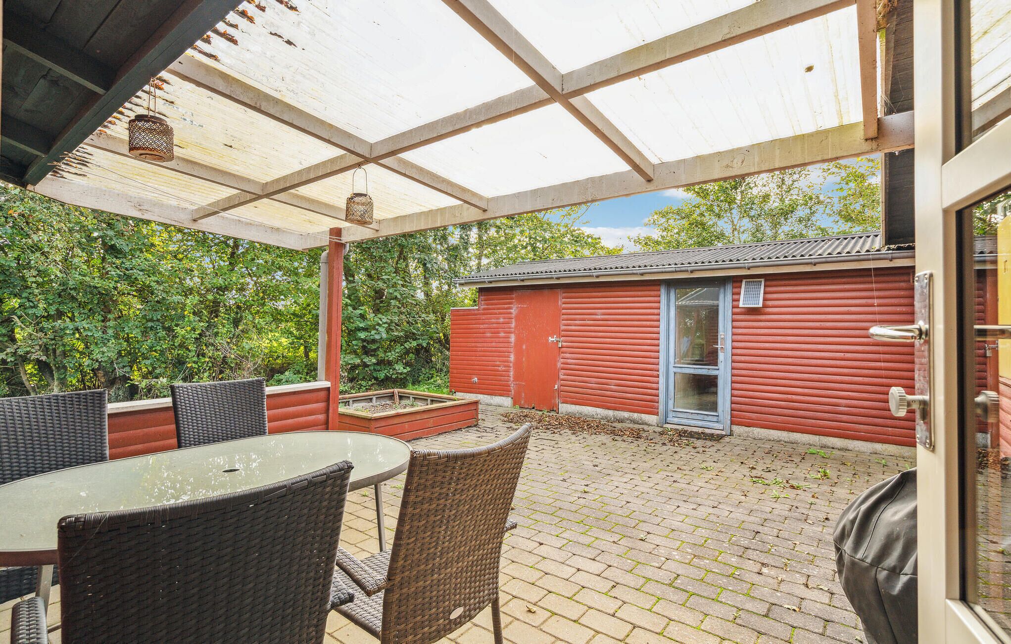 En overdækket terrasse med fletmøbler, en rød bygning med skydedør og et murstensgulv.