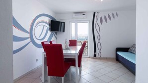 A dining area with a white table and red chairs, a flat-screen TV on the wall, and a balcony with a view.