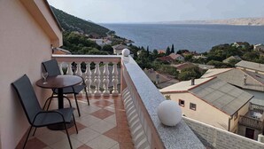 A balcony with a table and chairs, overlooking a coastal town and the sea.