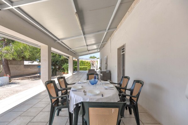 Une terrasse extérieure couverte, avec une table ronde dressée pour les repas, entourée de chaises, et offrant une vue sur le jardin.