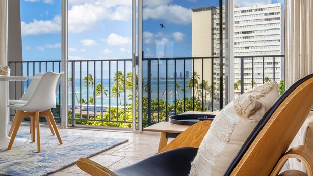 A balcony with a view of the ocean and palm trees, a dining area with a table and chairs, and a large window with a view of a cityscape.