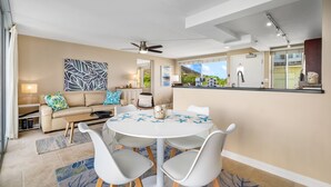 A modern living room with a round dining table, white chairs, a beige sofa with blue and green cushions, a ceiling fan, and a view of the outdoors through large windows.