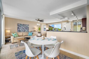 Uma sala de estar moderna com uma mesa de jantar redonda, cadeiras brancas, um sofá bege com almofadas azuis e verdes, um ventilador de teto e vista para o exterior através de grandes janelas.