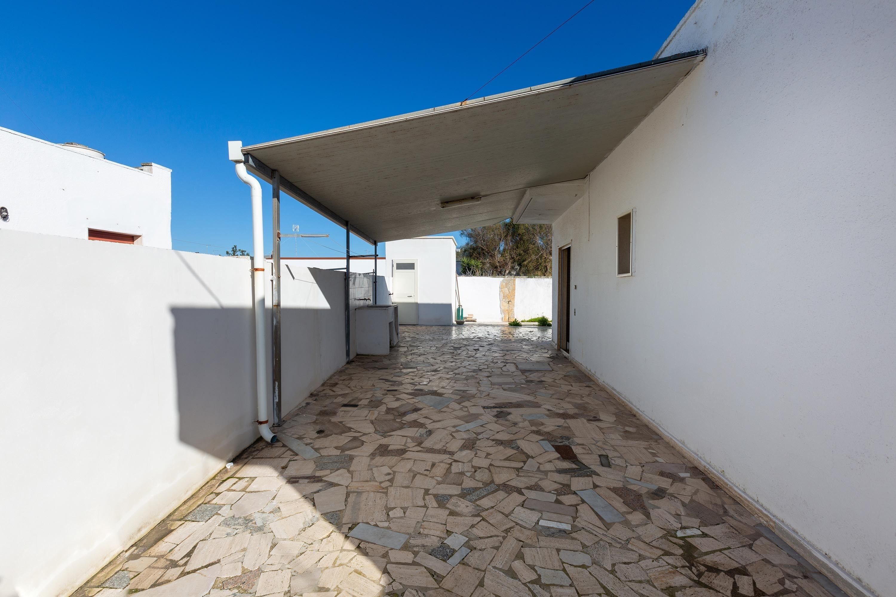 A narrow, tiled corridor with a covered walkway, white walls, and a clear blue sky.