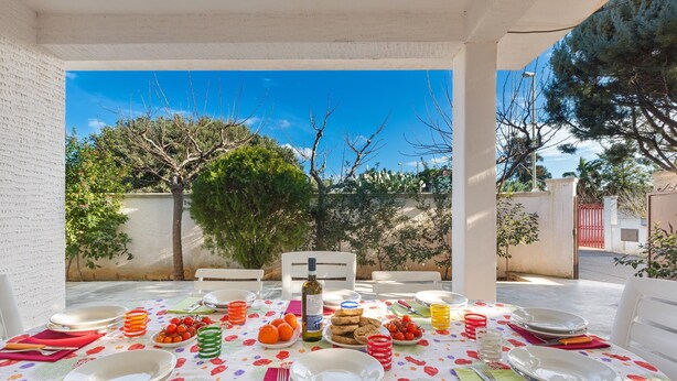 An outdoor dining area with a table set for a meal, surrounded by chairs, and a view of trees and a building in the background. - 2679 Villetta Filomena - Appartamento Leo by Barbarhouse (Manduria)