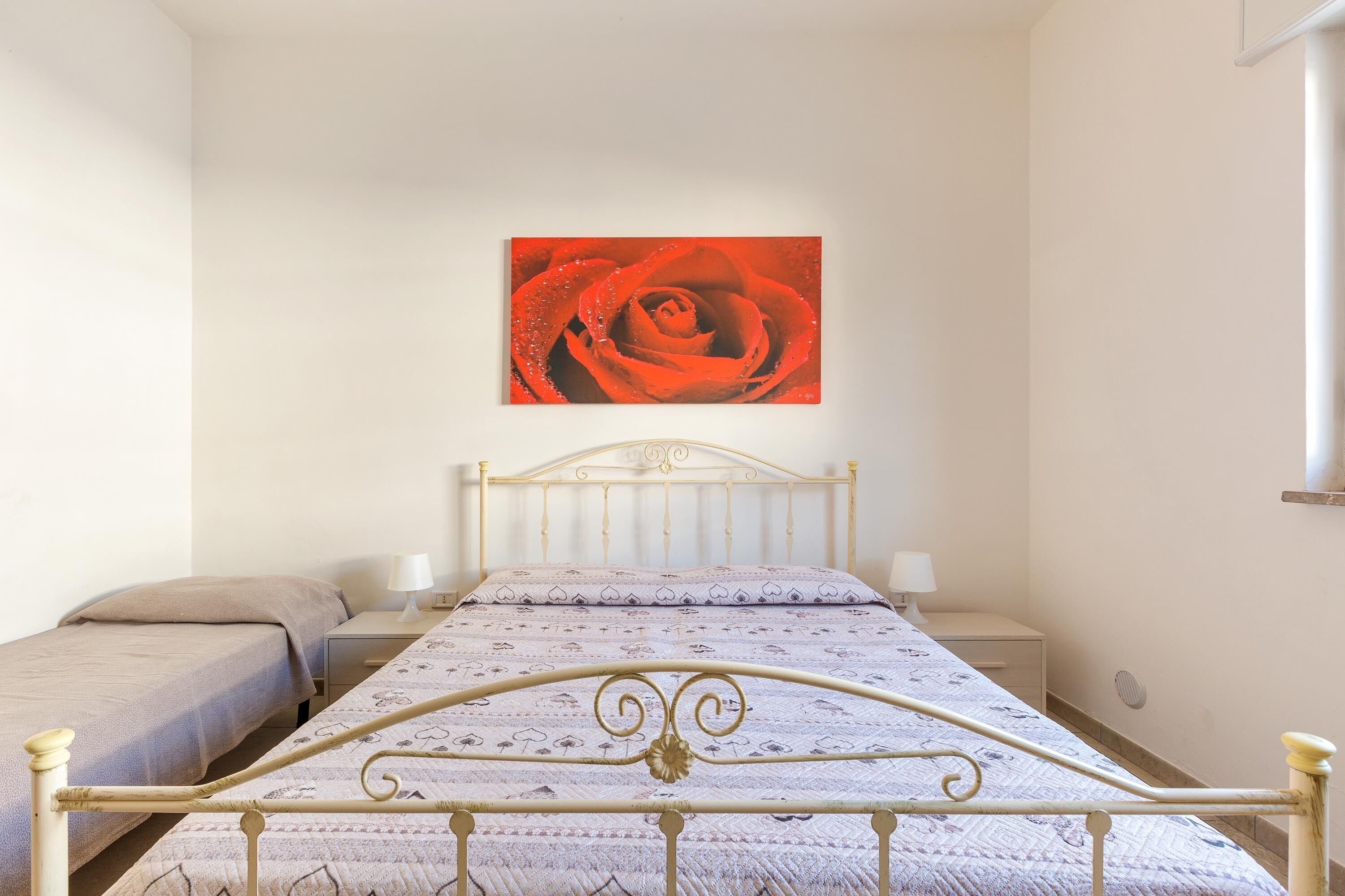 A bedroom with a metal bed frame, a single bed with a patterned bedspread, a bedside table with a lamp, and a wall-mounted painting of a rose.