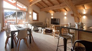 A spacious living area with a wooden ceiling, a large window offering a mountain view, a flat-screen TV, and a dining table with chairs. - Bain À Remous Privé! Chalet au coeur du Village (L'Alpe d'Huez)