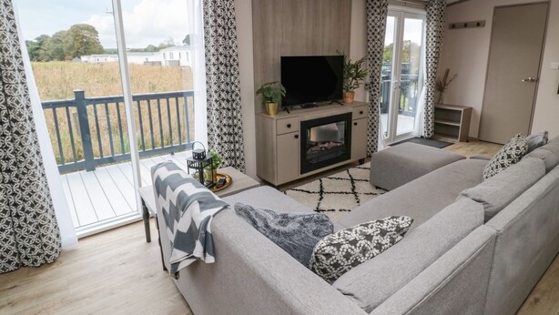 A modern living room with a sofa, a fireplace, a TV, and a balcony with patterned curtains. - Plot 18 Meadow View (Morpeth)