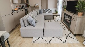A modern living room with a grey sectional sofa, a flat-screen TV, a fireplace, and a kitchen area with cabinets and a microwave. - Plot 18 Meadow View (Morpeth)