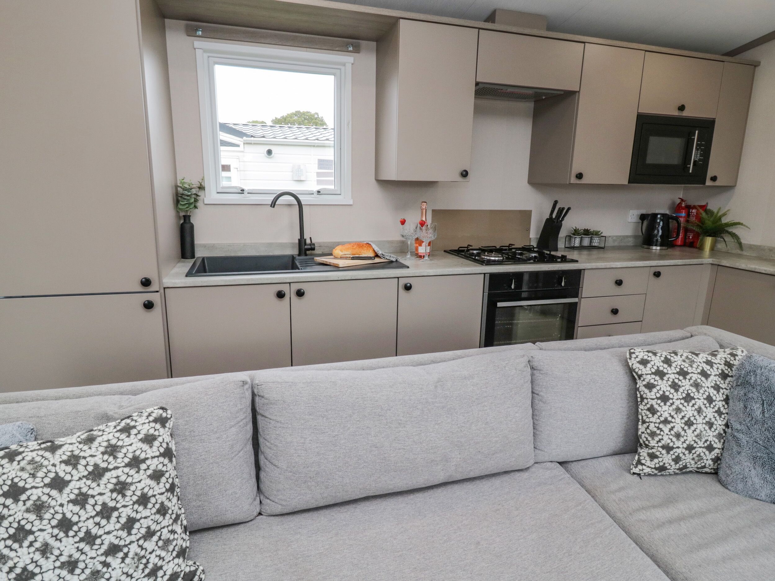 A compact kitchenette with a sink, stove, and microwave, a window with a view of a house, and a grey sofa with patterned cushions.